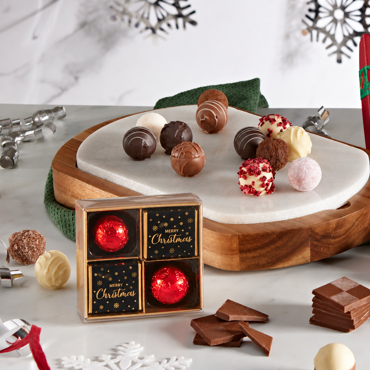 A boxed assortment of chocolates and truffles displayed on a marble board and tabletop, with holiday decorations in the background. The front box reads 'Merry Christmas' and contains four red-and-gold wrapped chocolates.