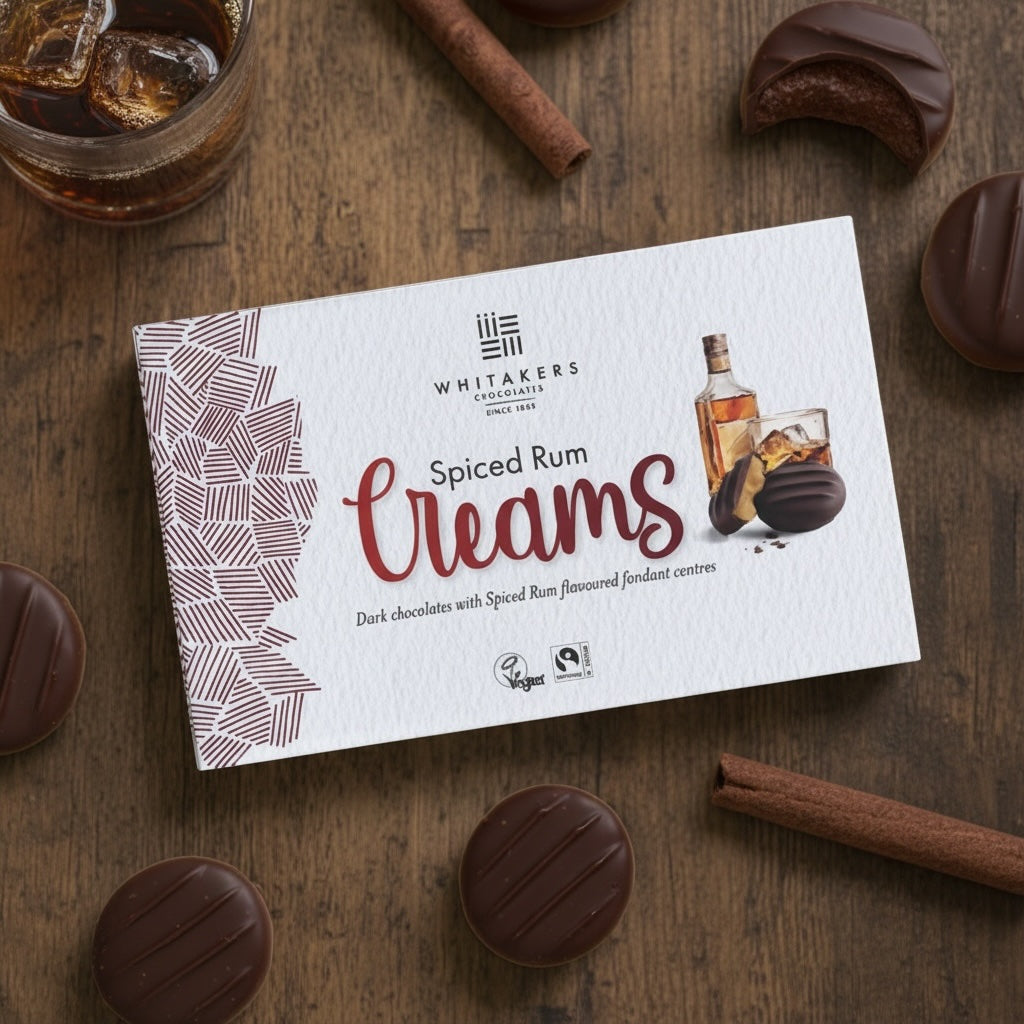 Box of Whitaker's dark chocolate Spiced Rum Creams on a wooden surface with chocolate candies and a glass of whiskey.