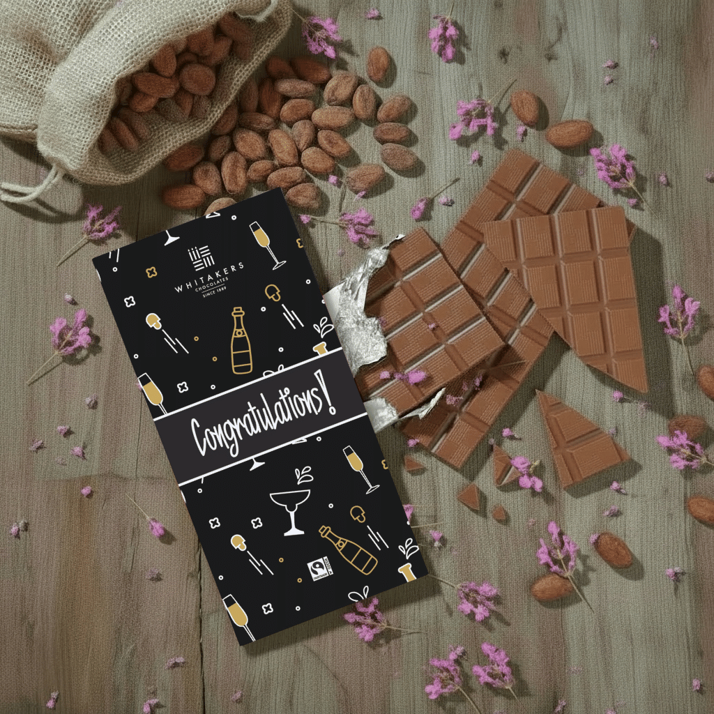 Whitaker's Congratulations milk chocolate bar on a rustic wooden background with cocoa beans, pink flowers and chocolate pieces.