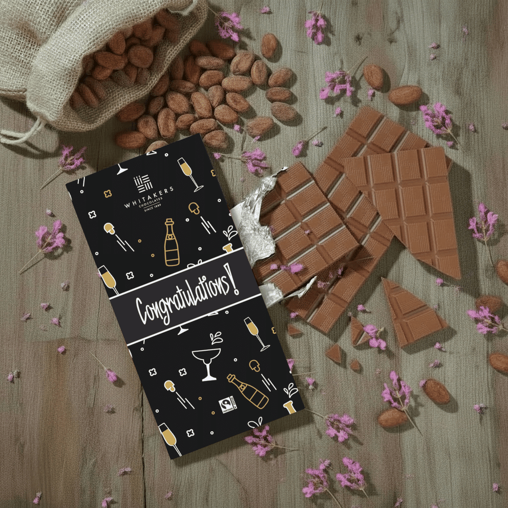 Whitaker's Congratulations milk chocolate bar on a rustic wooden background with cocoa beans, pink flowers and chocolate pieces.