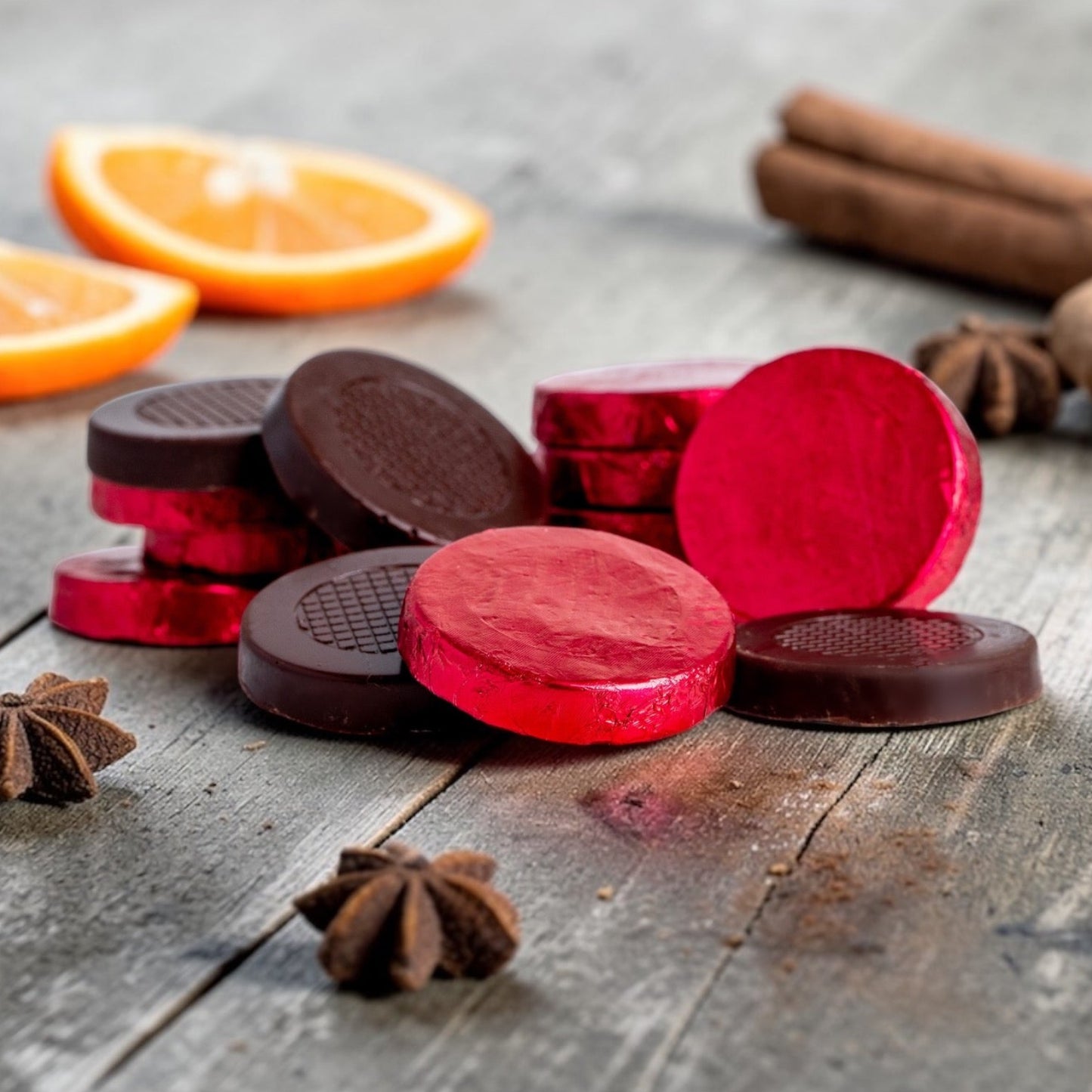 Stack of dark chocolate red foiled festive spice crisps on a rustic wooden background, with orange, cinnamon sticks and star anise scattered around the chocolates.