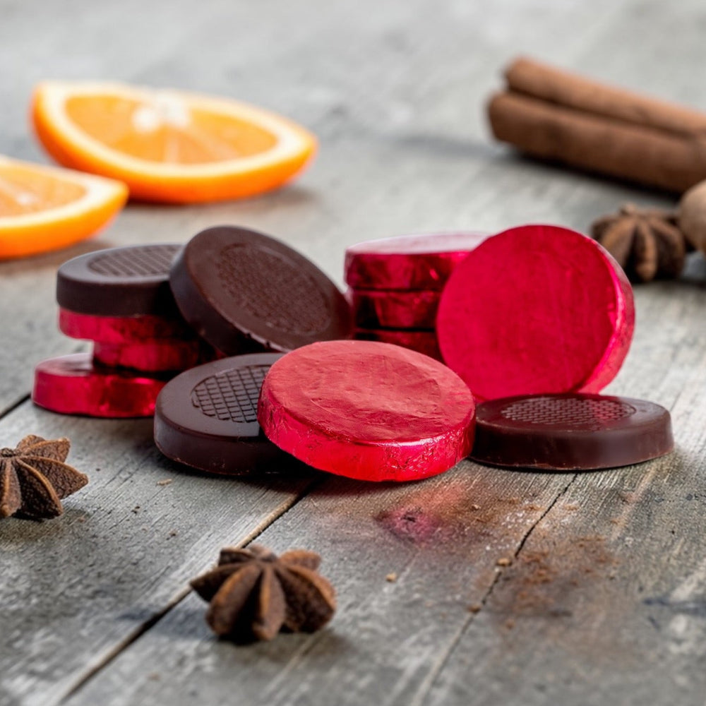 Stack of dark chocolate red foiled festive spice crisps on a rustic wooden background, with orange, cinnamon sticks and star anise scattered around the chocolates.