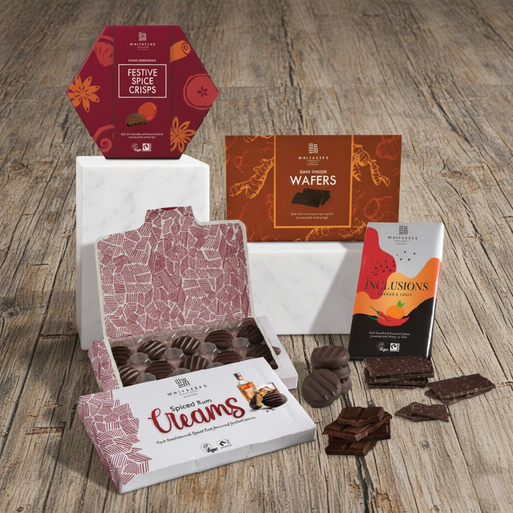 Whitakers Chocolates Assorted chocolate products including Fairtrade 'Festive Spice Crisps', 'ginger wafers', 'Chilli and orange Inclusions bar', and 'spiced rum Creams' on a wooden surface.