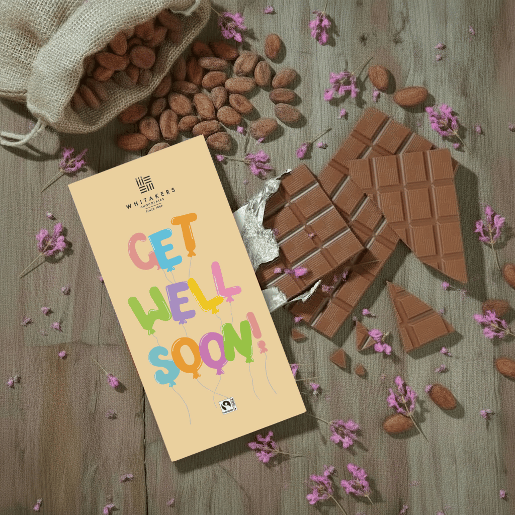 Whitaker's Get Well Soon milk chocolate bar on a rustic wooden background with cocoa beans, pink flowers and chocolate pieces.