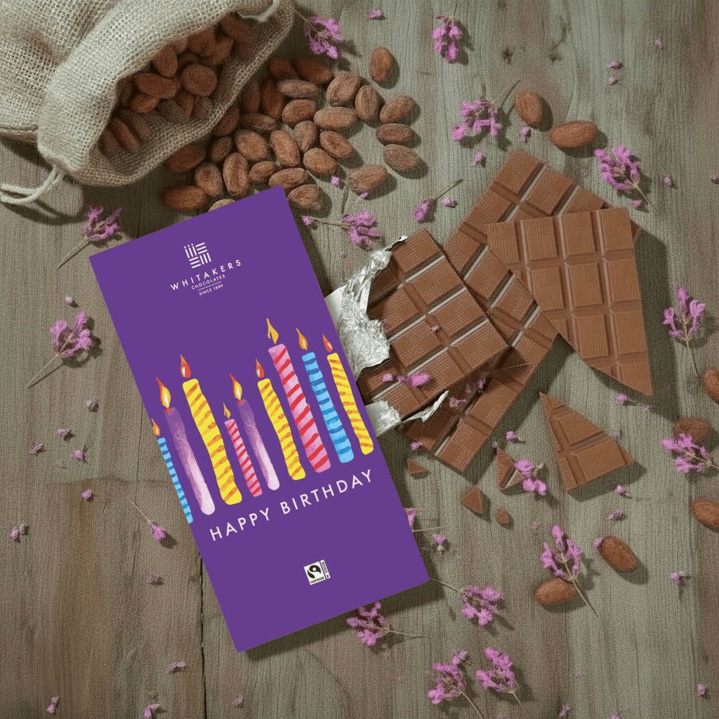 Whitaker's Happy Birthday Candles milk chocolate bar on a rustic wooden background with cocoa beans, pink flowers and chocolate pieces.