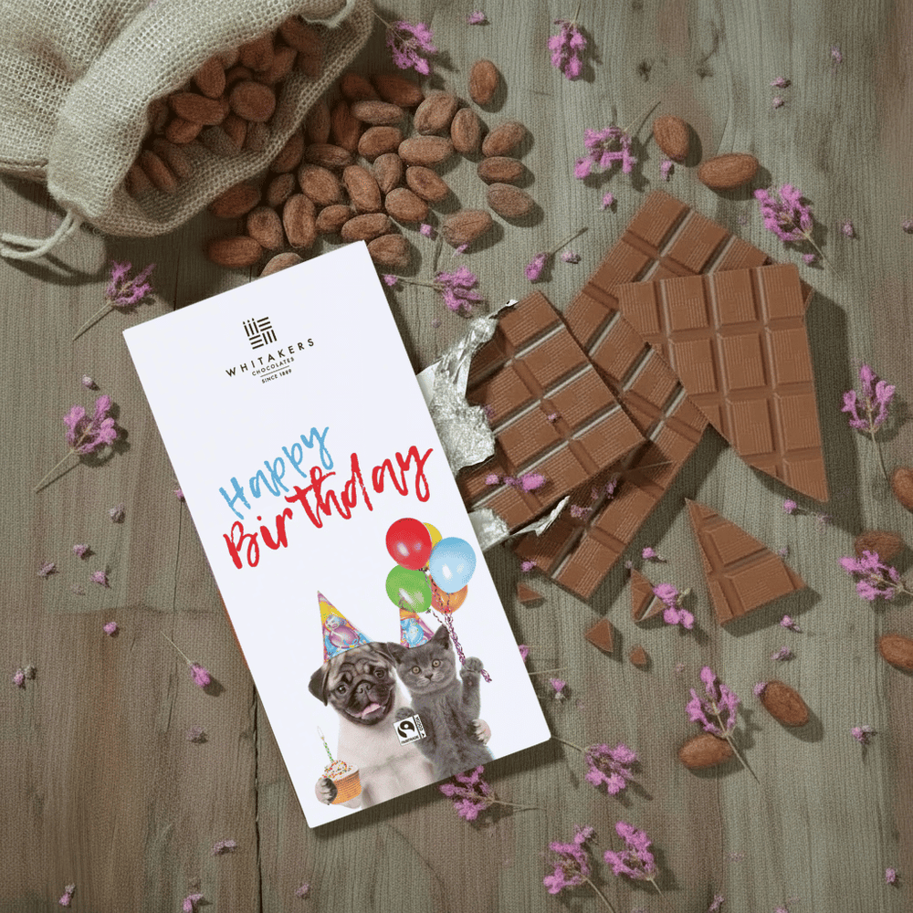 Whitaker's Happy Birthday from the Pets milk chocolate bar on a rustic wooden background with cocoa beans, pink flowers and chocolate pieces.