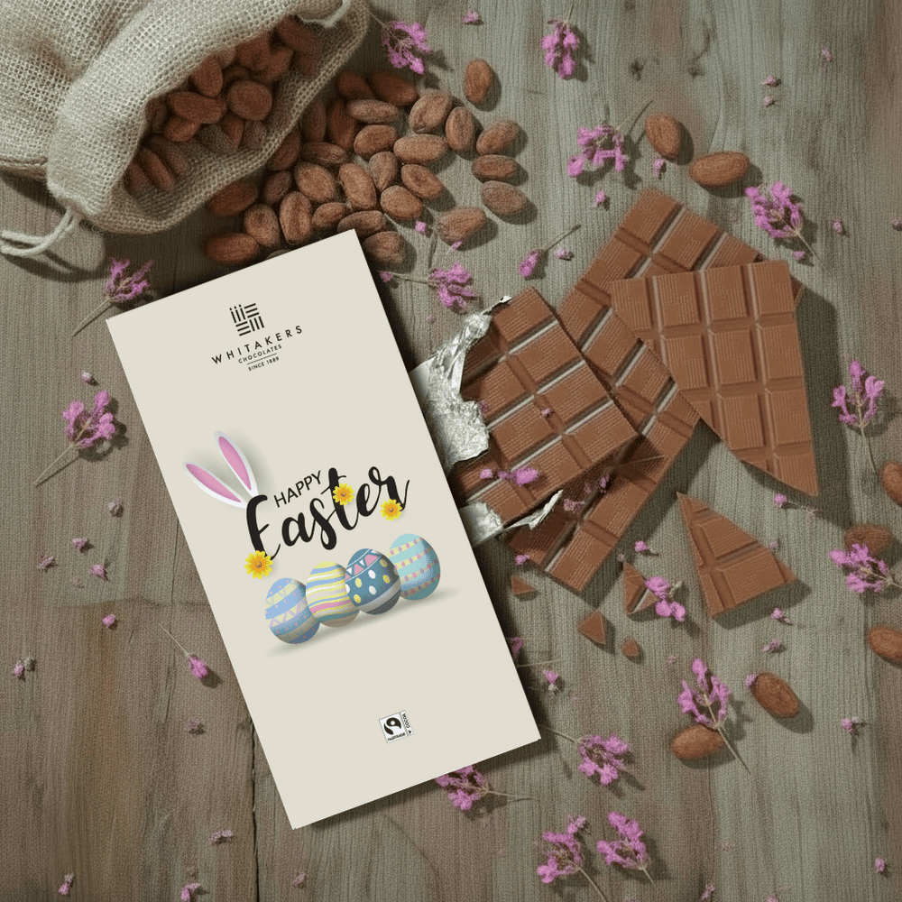 Whitaker's Happy Easter (eggs) milk chocolate bar on a rustic wooden background with cocoa beans, pink flowers and chocolate pieces.