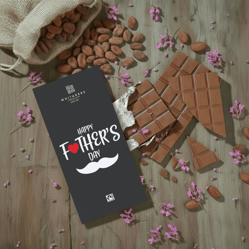 Whitaker's Happy Fathers Day milk chocolate bar on a rustic wooden background with cocoa beans, pink flowers and chocolate pieces.