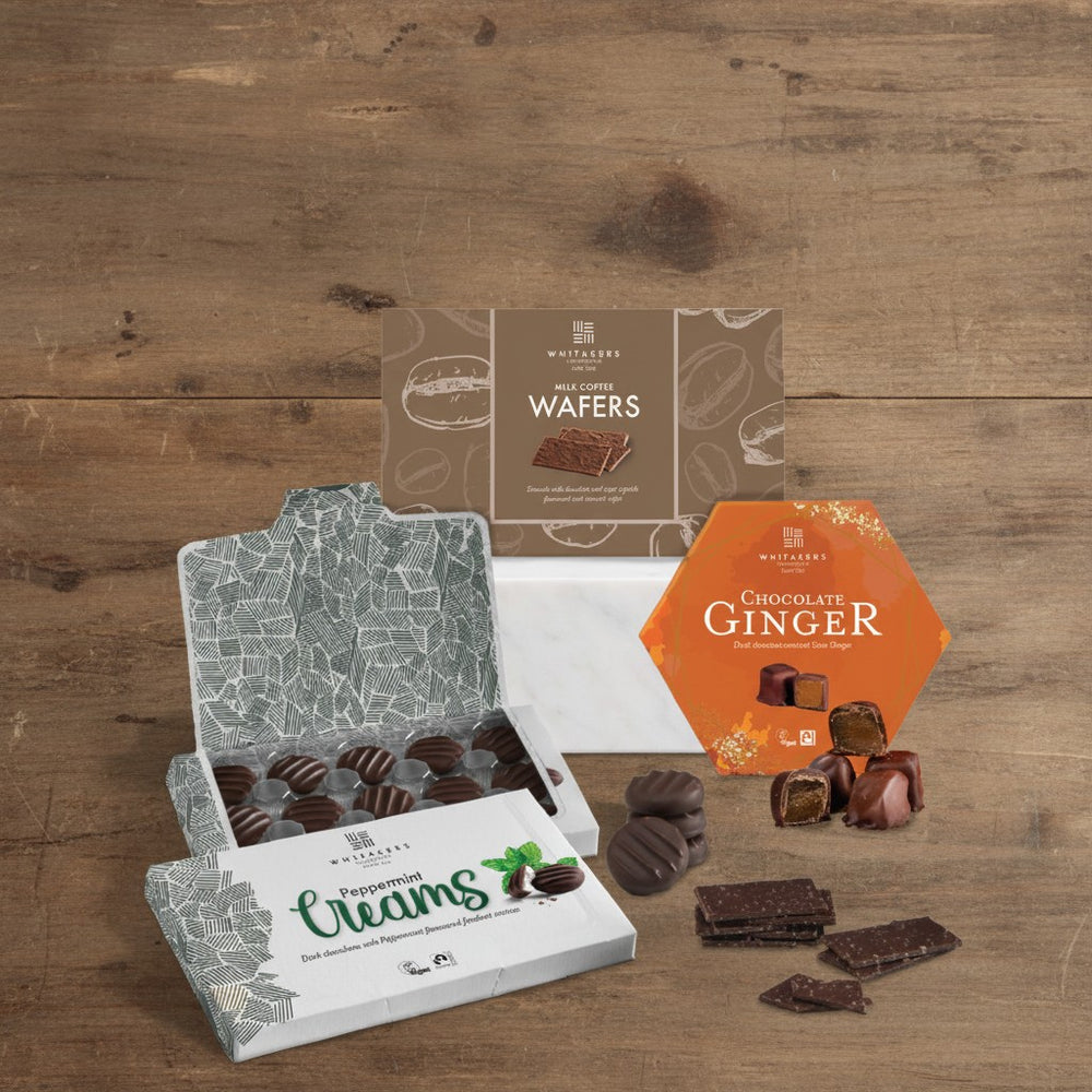 Whitakers Chocolates Assorted chocolate products including milk coffee wafers, dark stem ginger, and peppermint creams on a wooden surface.