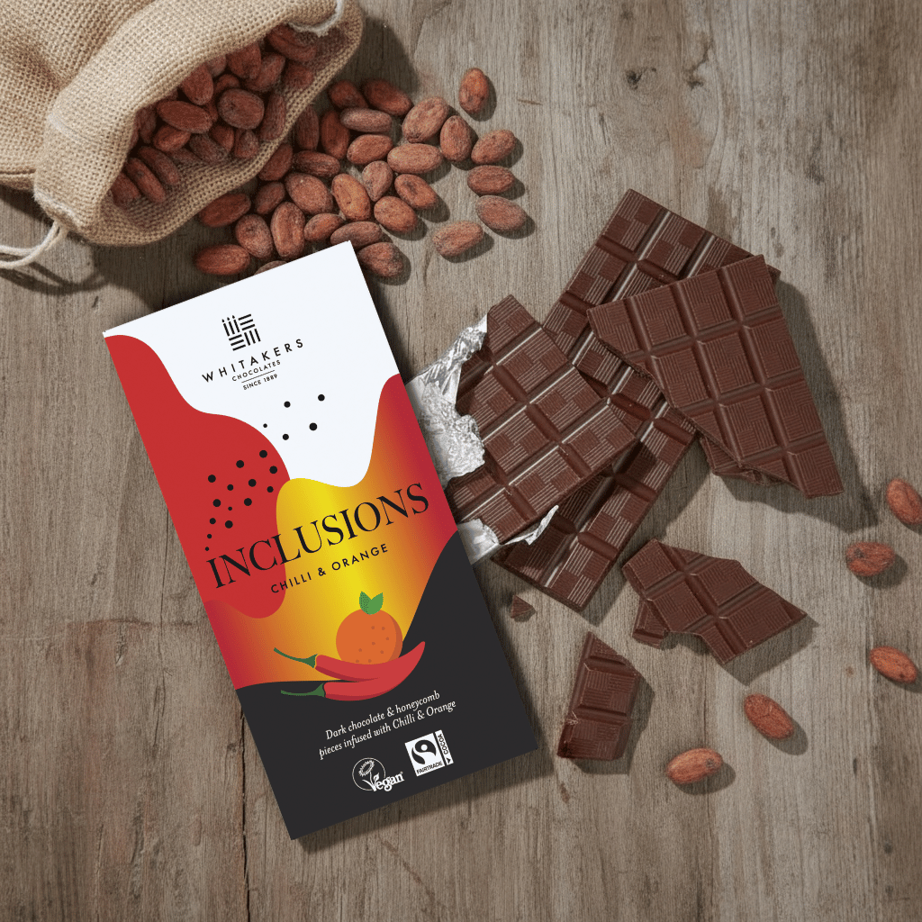 Whitaker's Dark chocolate Chilli and Orange 90g bar on a rustic wooden surface with loose cocoa beans and chocolate pieces