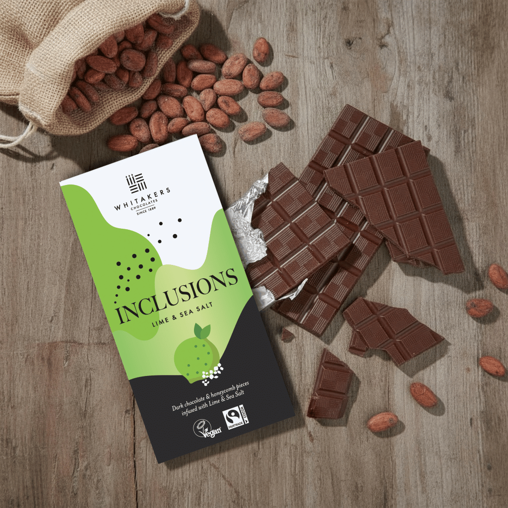 Whitaker's Dark chocolate Lime and Sea Salt 90g bar on a rustic wooden surface with loose cocoa beans and chocolate pieces