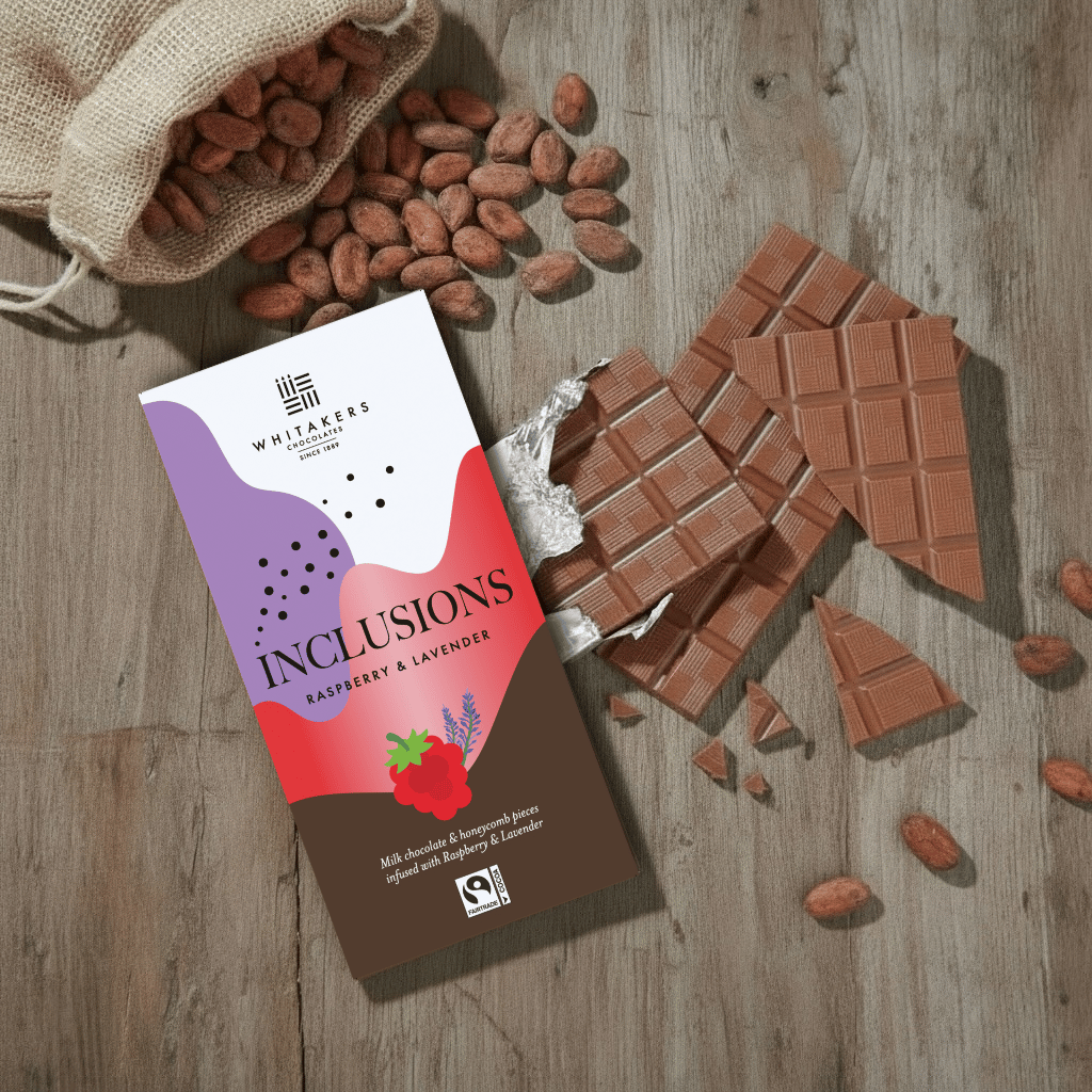 Whitaker's Milk chocolate Raspberry and Lavender 90g bar on a rustic wooden surface with loose cocoa beans and chocolate pieces