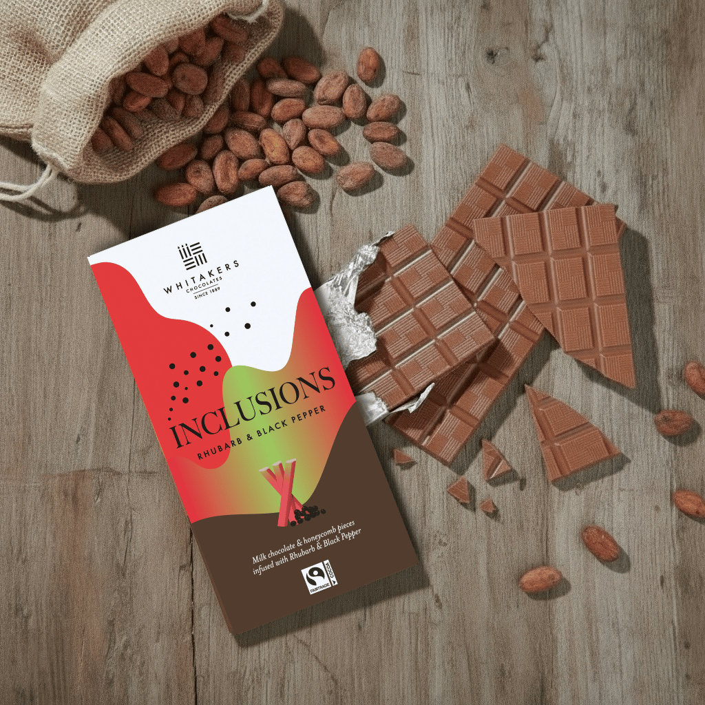 Whitaker's Milk chocolate Rhubarb adn black pepper 90g bar on a rustic wooden surface with loose cocoa beans and chocolate pieces