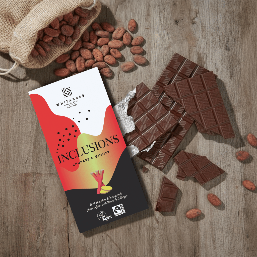 Whitaker's Dark chocolate Rhubarb and Ginger 90g bar on a rustic wooden surface with loose cocoa beans and chocolate pieces