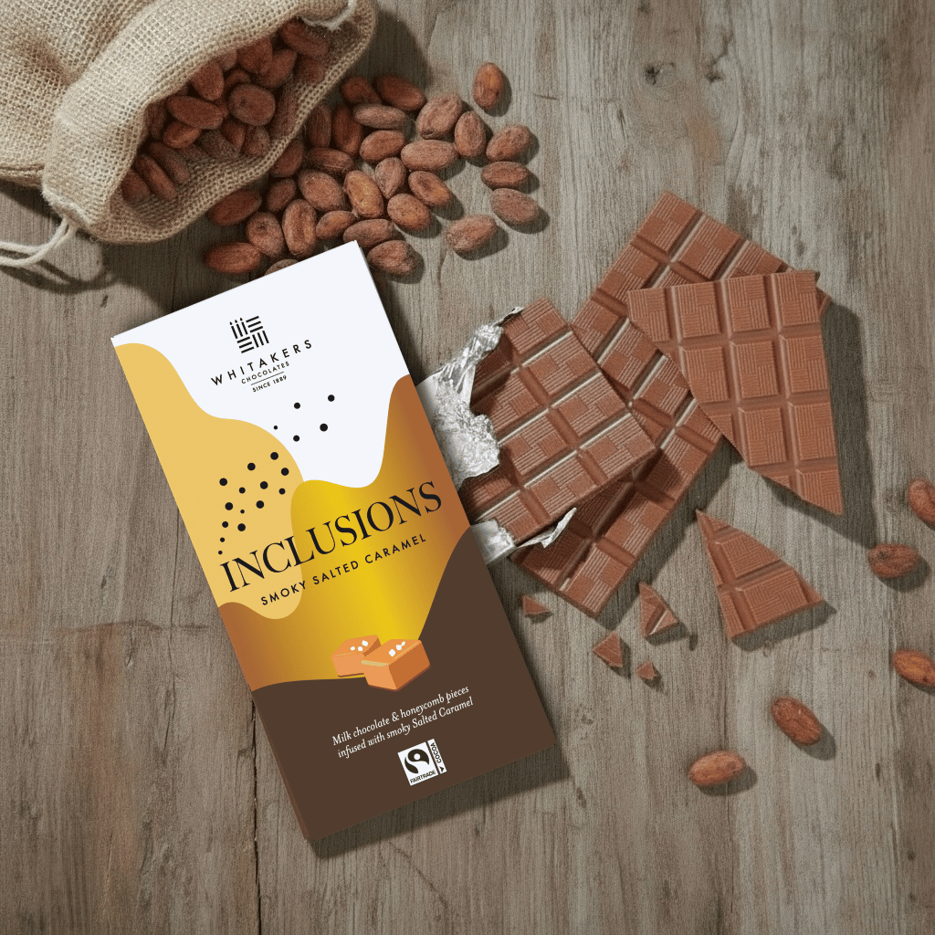 Whitaker's Milk chocolate smoky salted caramel 90g bar on a rustic wooden surface with loose cocoa beans and chocolate pieces