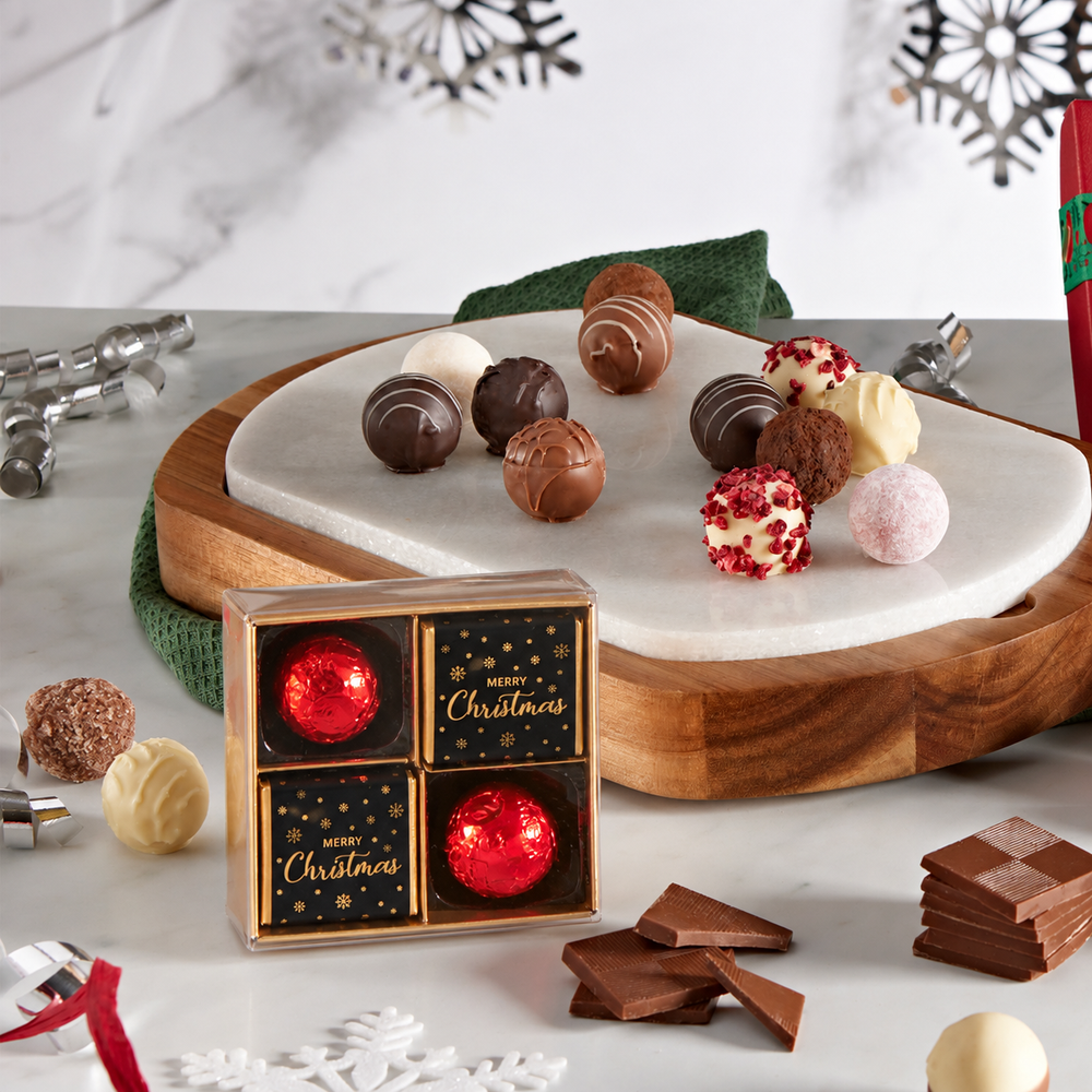 A boxed assortment of chocolates and truffles displayed on a marble board and tabletop, with holiday decorations in the background. The front box reads 'Merry Christmas' and contains four red-and-gold wrapped chocolates.