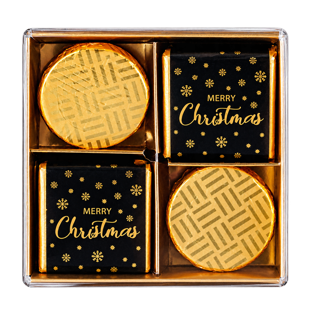 A boxed set of four holiday chocolates in a clear square gift box. Two chocolates are wrapped in shiny gold foil, and two are wrapped in black paper with gold 'MERRY Christmas' text and snowflake motifs.