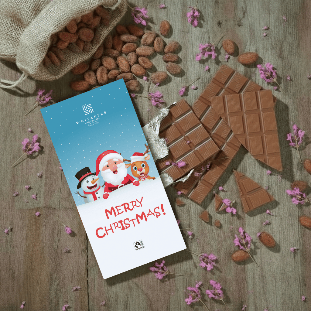Whitaker's Merry Christmas Cartoon milk chocolate bar on a rustic wooden background with cocoa beans, pink flowers and chocolate pieces.