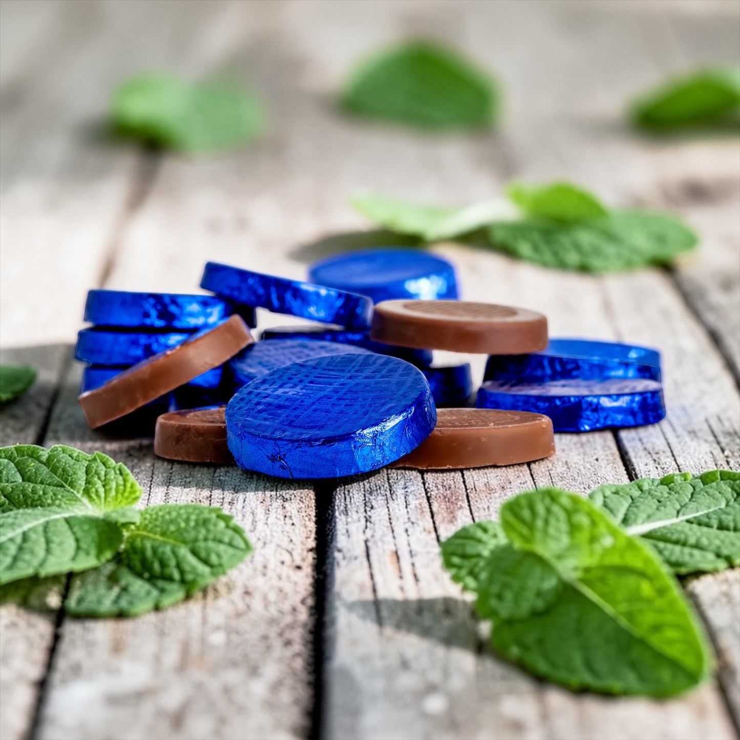 Stack of milk chocolate mint crisps in blue foil, on a rustic wooden background with mint leaves scattered in the background and foreground