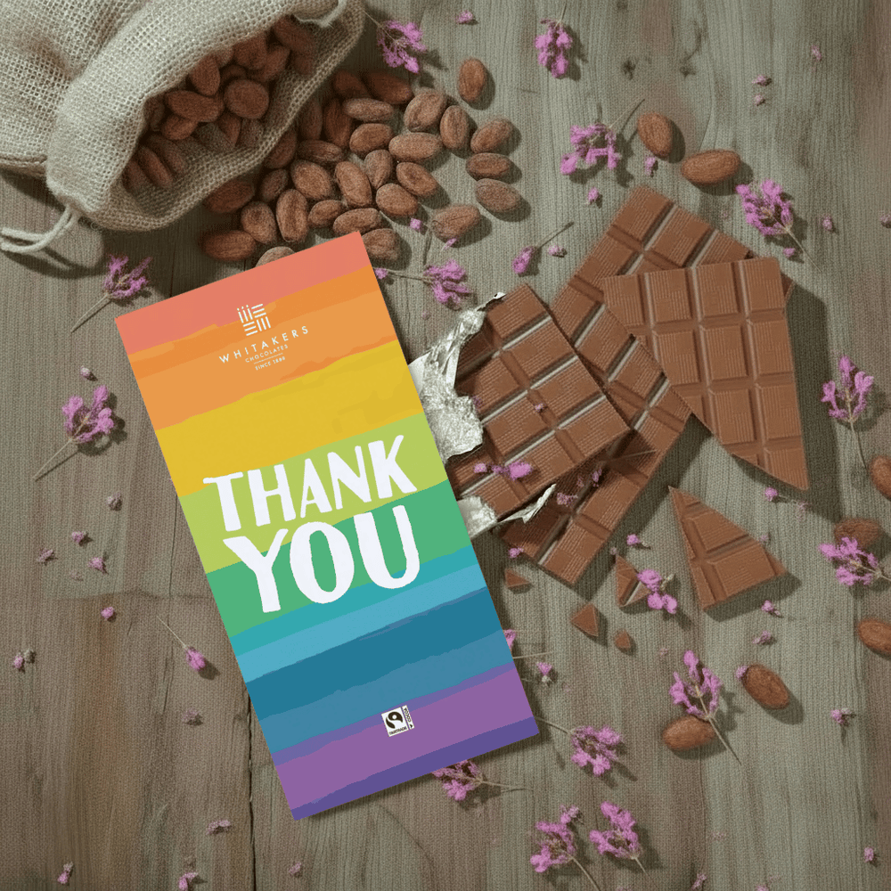 Whitaker's Rainbow Thank You milk chocolate bar on a rustic wooden background with cocoa beans, pink flowers and chocolate pieces.