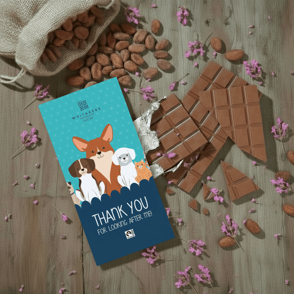 Whitaker's Thank You For Looking After me (pets) milk chocolate bar on a rustic wooden background with cocoa beans, pink flowers and chocolate pieces.