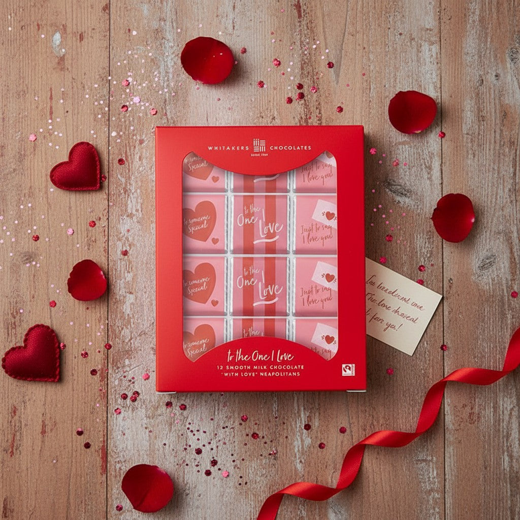 Box of chocolates with heart designs on a wooden surface with red ribbons and petals.