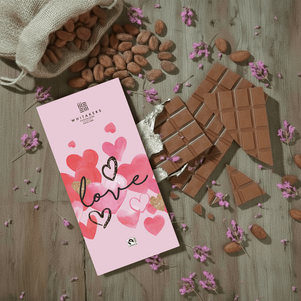 Whitaker's Valentines Love milk chocolate bar on a rustic wooden background with cocoa beans, pink flowers and chocolate pieces.