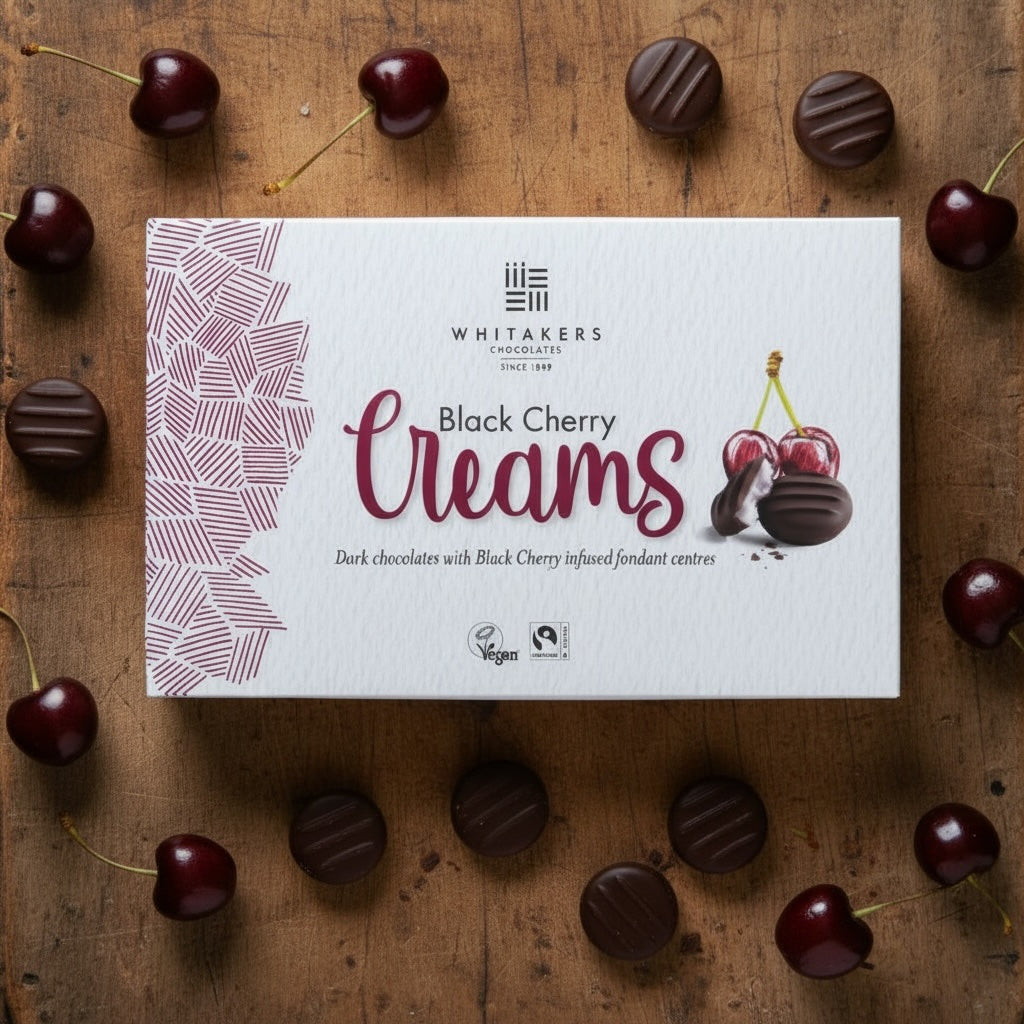 Box of Whitaker's dark chocolate Black Cherry Creams on a wooden surface with cherries and chocolates.