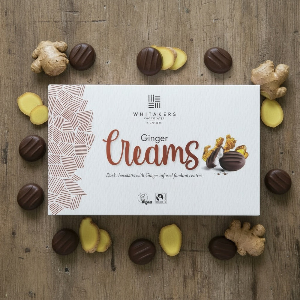 Box of Whitaker's dark chocolate Ginger Creams chocolates on a wooden surface with ginger pieces.