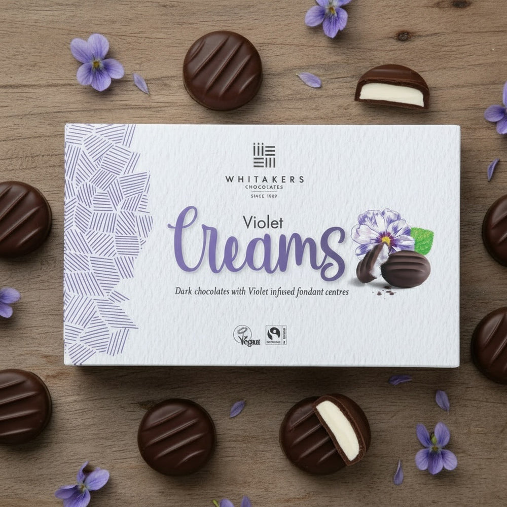 Box of Whitaker's Violet Creams chocolates on a wooden surface with flowers.