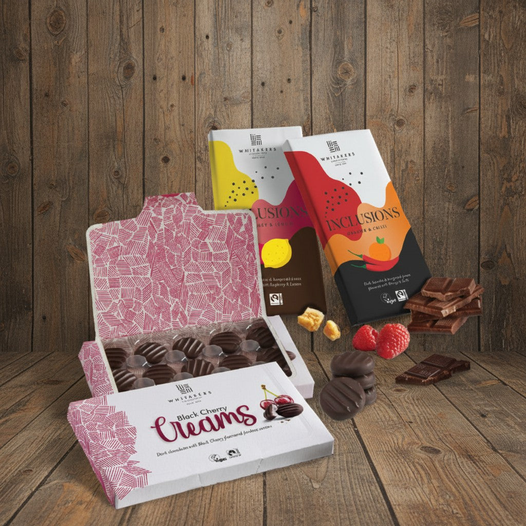 Whitakers Fairtrade Chocolate Lovers Bundle, containing dark chocolate black cherry creams, dark chocolate chill & orange bar, raspberry & lemon milk chocolate bar on a rustic wooden surface