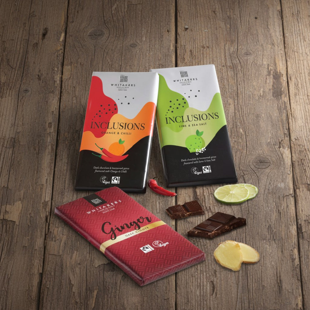 Three Whitakers Chocolates dark chocolate bars on a wooden surface with ingredients visible, bar flavours are chilli & orange, lime and sea salt and ginger