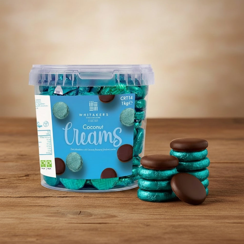 Container of Whitakers dark chocolate turquoise foiled coconut creams with a stack of chocolates on a wooden surface